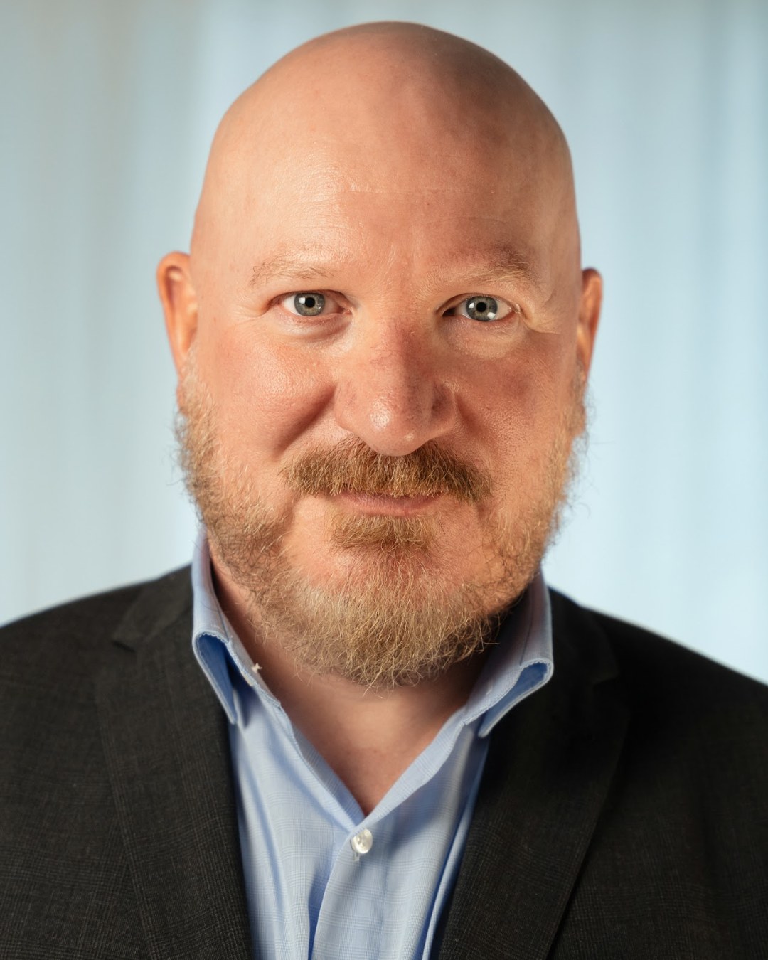 Portrait de Roland Sheppard, formateur en relation client et gestion des plaintes pour l’hôtellerie et le retail premium, souriant, en veste sombre et chemise bleu ciel